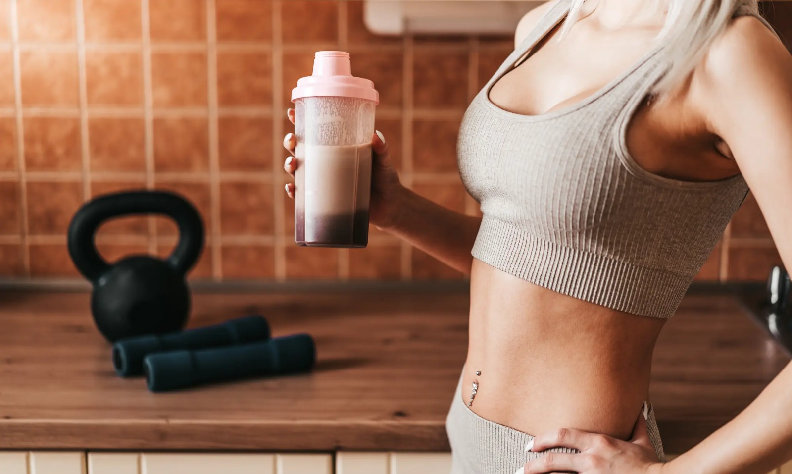 Woman in light beige sports clothing holding a protein shake with workout equipment in the background